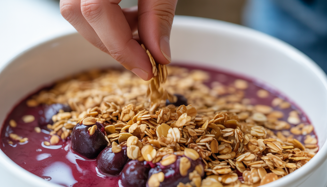 A hand sprinkling granola over an acai bowl, focusing on the rich purple acai and golden brown granola, showcasing customization in healthy meals.